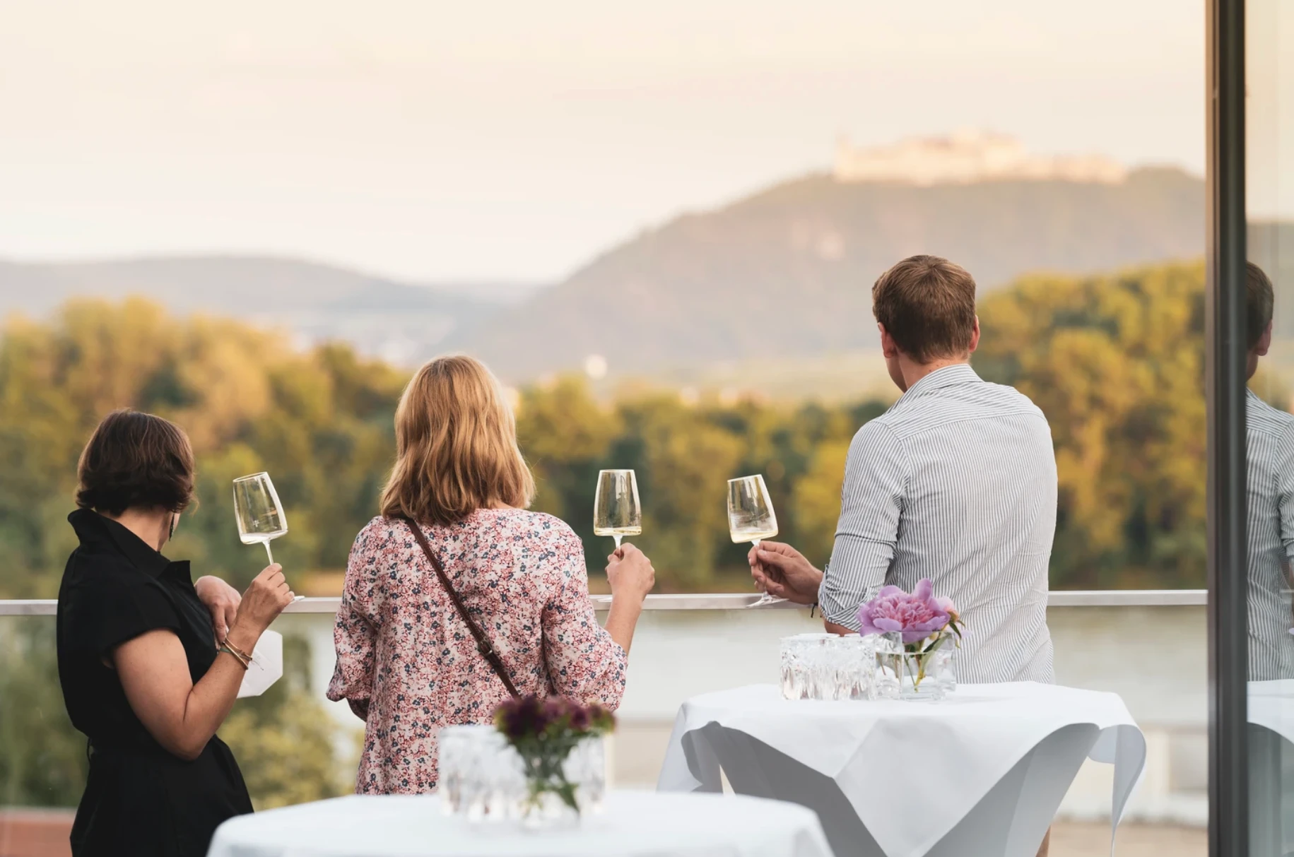 Drei Personen geniessen Weisswein auf einem Balkon mit Blick auf eine Flusslandschaft und ein Schloss